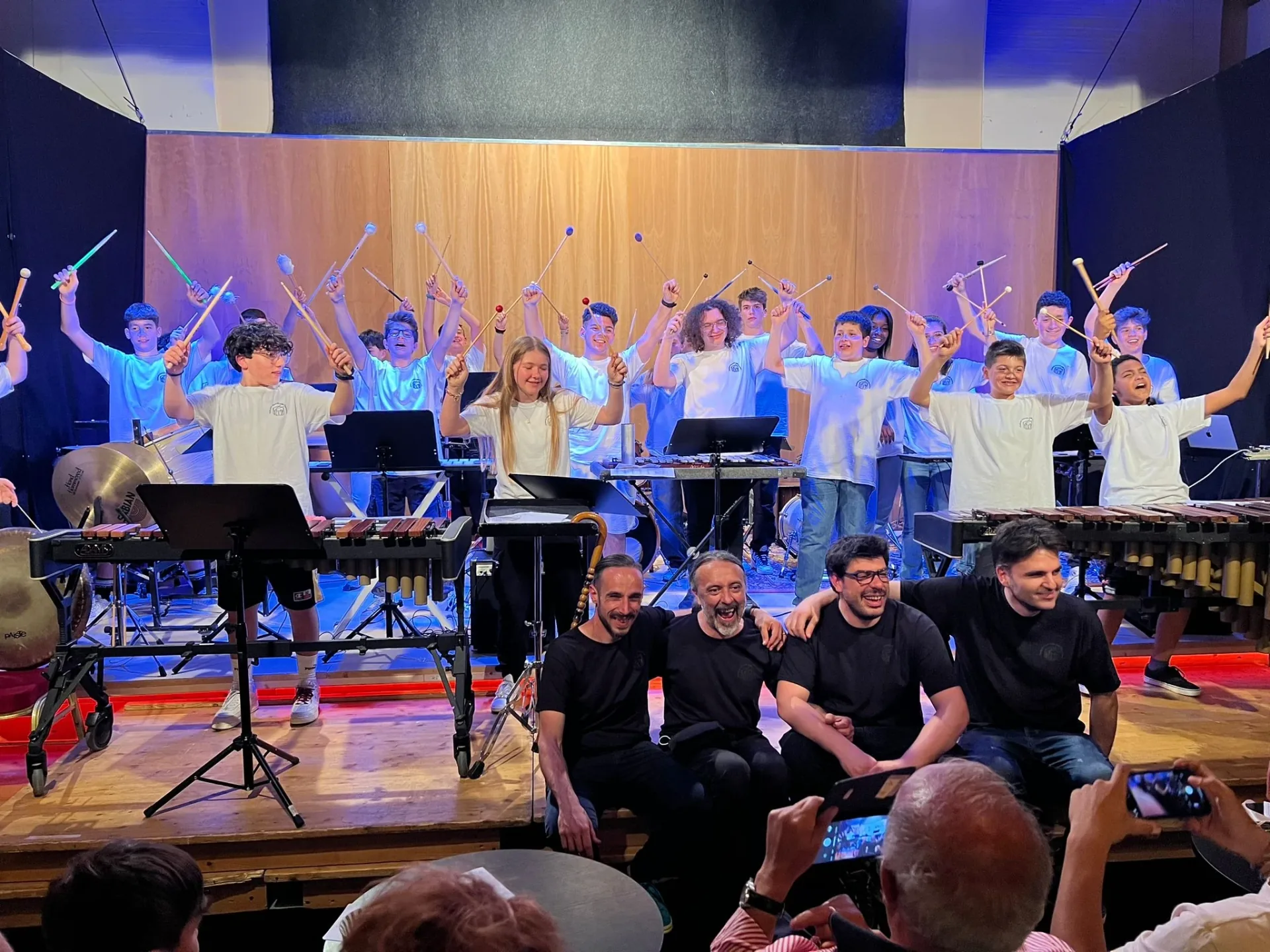 Foto di gruppo a fine concerto: i ragazzi sul palco con le bacchette alzate e lo staff seduto davanti